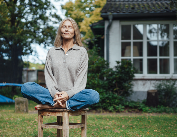 Eine Frau sitzt im Schneidersitz auf einem Holzstuhl im Garten und meditiert