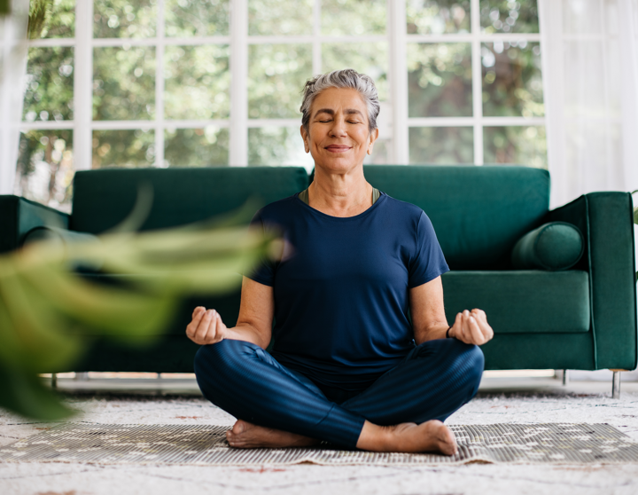 Eine ältere Dame sitzt in ihrem Wohnzimmer auf ihrer Yoga Matte und meditiert.