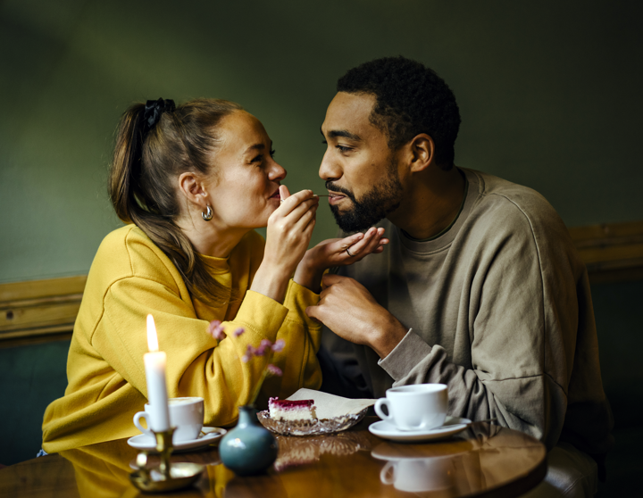 Ein junges Paar sitzt entspannt an einem kleinen Essenstisch und die Frau führt ein Stück Kuchen zu dem Mund von dem Mann