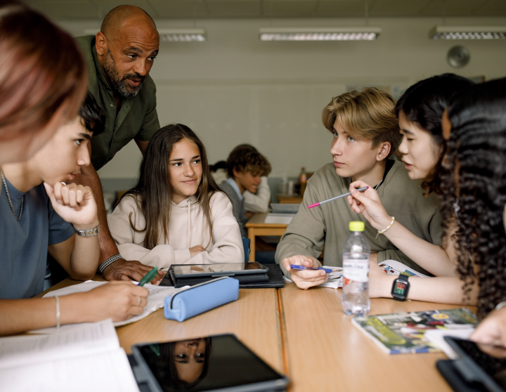 Ein paar Schüler und Schülerinnen sitzen gemeinsam an einem Tisch und der Lehrer erklärt ihnen ein Finanzthema