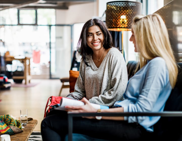 Eine brünette und eine blonde Frau sitzen auf einem Sofa in einem Café und reden und lächeln sich an