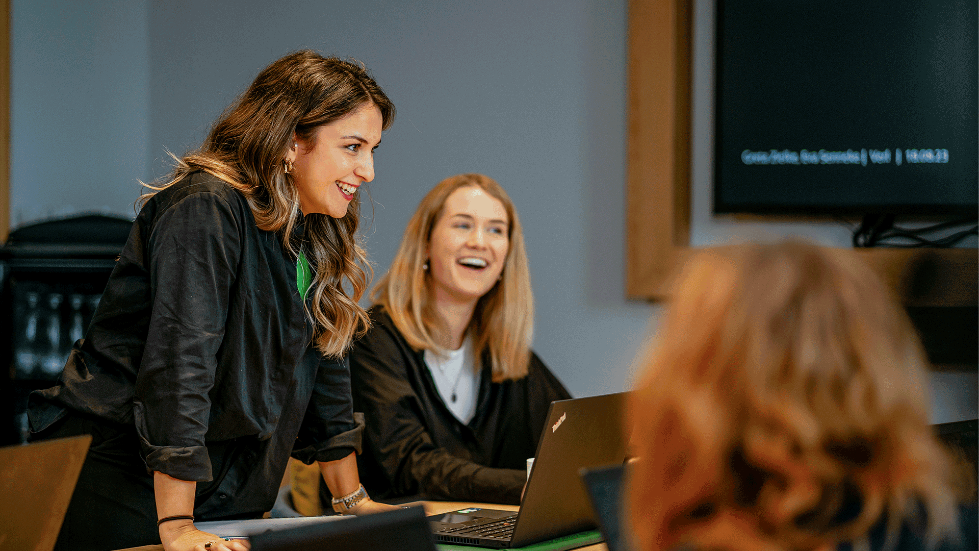 Zwei lächelnde Frauen in einem Büro, die zusammen an einem Laptop arbeiten. Eine sitzt, die andere steht neben ihr und zeigt auf den Bildschirm, während eine dritte Person mit dem Rücken zur Kamera sitzt.