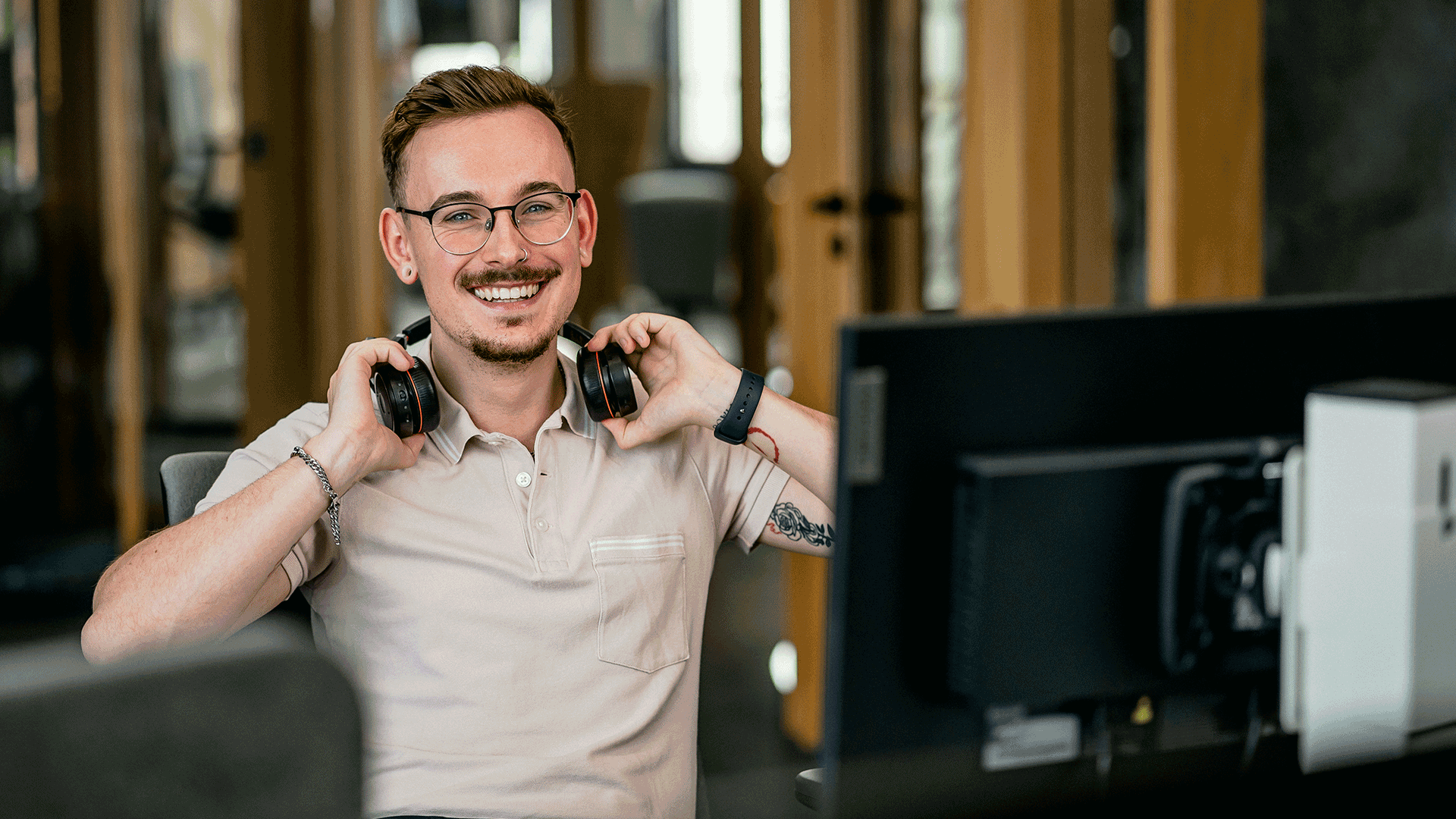 Ein junger Mann mit Brille und einem lächelnden Gesichtsausdruck sitzt an einem Schreibtisch in einem Büro. Er hält ein Paar Kopfhörer um seinen Hals und trägt ein hellbeiges Hemd. Am Handgelenk trägt er eine Smartwatch und am Unterarm ist ein Tattoo sichtbar. Vor ihm steht ein Computermonitor, und im Hintergrund sind Bürofenster, die für eine helle und freundliche Atmosphäre sorgen.