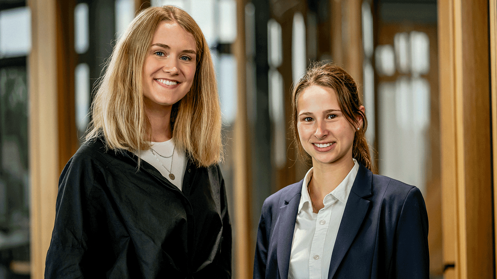 Zwei junge Frauen, die professionell gekleidet sind und in einem Bürokorridor stehen. Sie lächeln und blicken direkt in die Kamera. Die Frau auf der linken Seite trägt eine schwarze Jacke über einem weißen Oberteil und eine Halskette, während die Frau auf der rechten Seite ein dunkelblaues Jackett über einer weißen Bluse trägt. Der Hintergrund ist unscharf, mit großen Fenstern und Türen, die natürliches Licht hereinlassen.