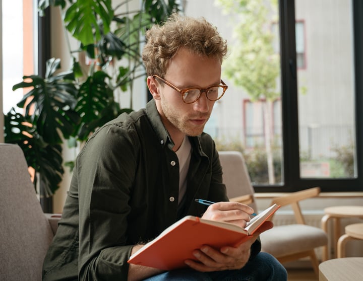 A man with glasses sits on an armchair and writes in a red book