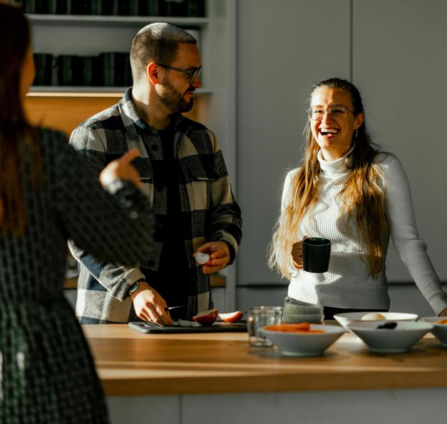 Three people are chatting and laughing in a modern kitchen. One person with long brown hair is standing with their back to the camera, gesturing. Another person with glasses, a beard, and a checkered shirt is cutting fruit and looking towards the first person. The third person, with long hair, glasses, and a white turtleneck sweater, leans against the kitchen island, holding a black mug and laughing. On the countertop, there are bowls of fruit and vegetables, as well as pots with fresh herbs. Sunlight streams through the window, illuminating the scene.