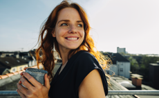 Eine glückliche junge Frau steht auf ihrem Balkon und hält eine Tasse Kaffee in der Hand