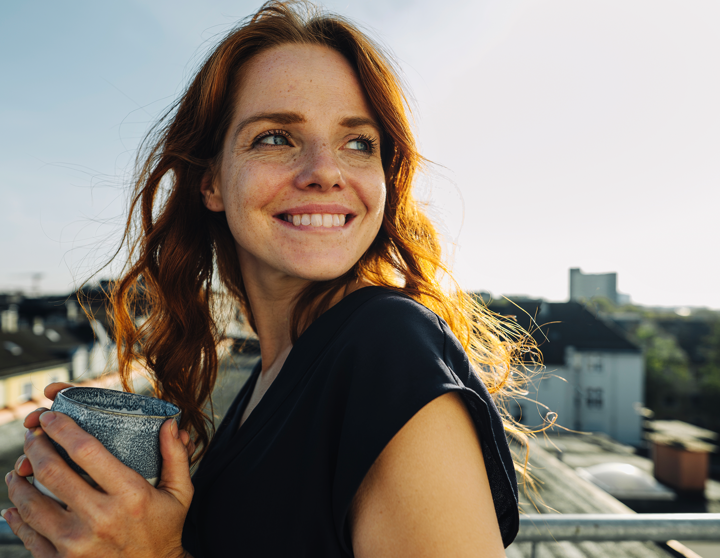 Eine glückliche junge Frau steht auf ihrem Balkon und hält eine Tasse Kaffee in der Hand