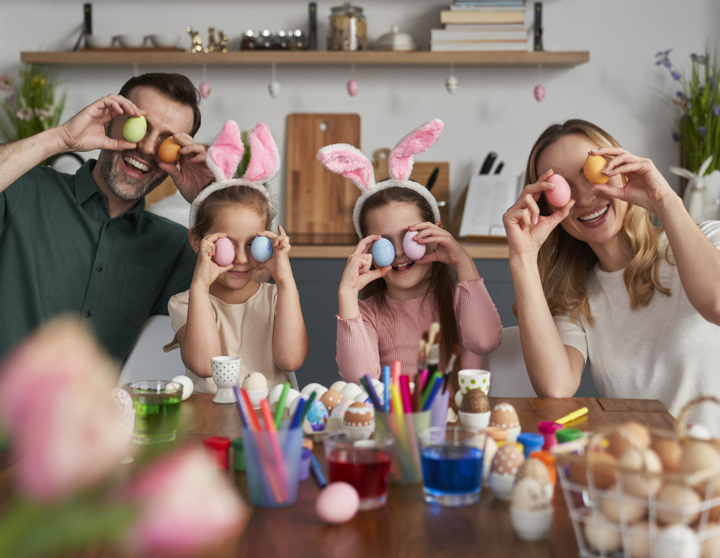 Eine glückliche Familie bemalt Ostereier und hält sich die bunten Ostereier als Augen vor ihre Gesichter