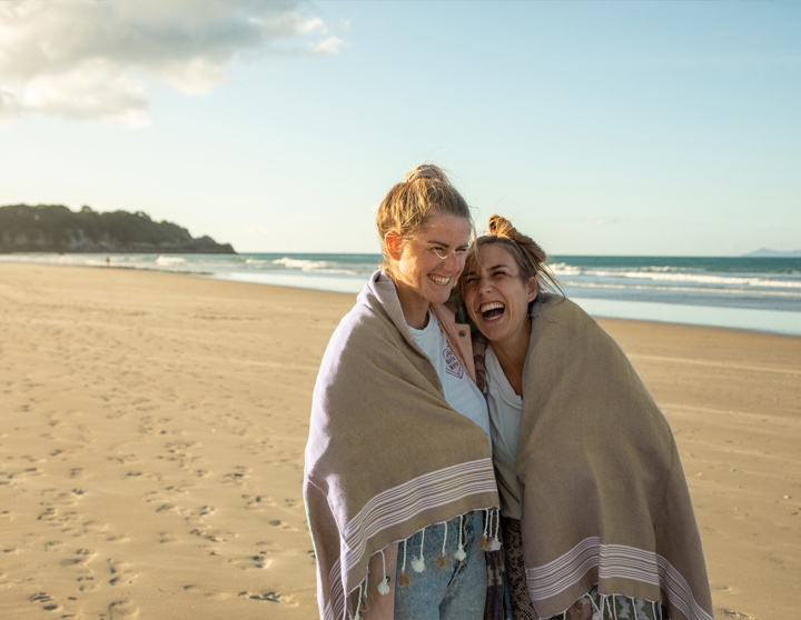 Zwei junge Frauen laufen mit einer Decke um ihre Schultern lachend am Strand