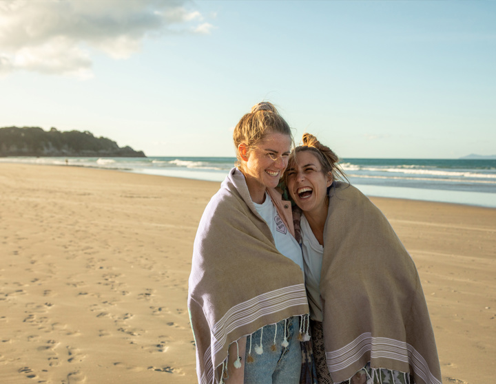 Twee jonge vrouwen lopen lachend over het strand met een deken om hun schouders