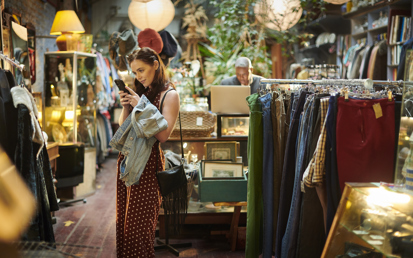 Eine brünette Frau steht mitten in einem Second-Hand-Geschäft und schaut lächelnd auf ihr Handy
