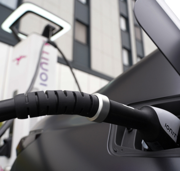 Close-up view of an electric vehicle being charged at an Ionity charging station, with the charging plug inserted into the car's port and part of the charging unit visible in the background.