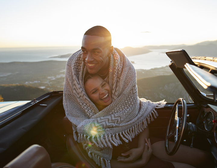 Eine junge Frau sitzt in einem Cabrio und ein junger Mann umarmt sie mit einer Decke von hinten