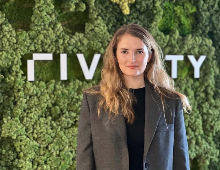 Anna steht vor einer Wand, die mit grünem Moos bedeckt ist. Hinter ihrem Kopf sieht erkennt man das Riverty Logo.