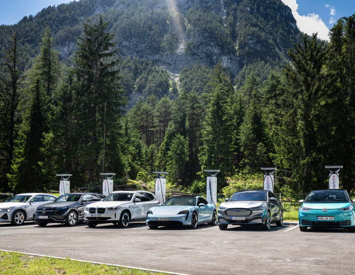 Six electric vehicles from various manufacturers are parked side by side at IONITY charging stations in a scenic mountainous area surrounded by dense pine forest and steep cliffs under a partly cloudy sky