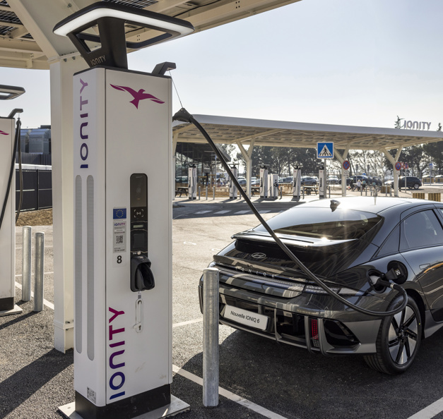 A black Hyundai Ioniq 6 electric vehicle is plugged into an Ionity fast-charging station at a modern EV charging facility, with several other chargers and a covered canopy visible in the background.