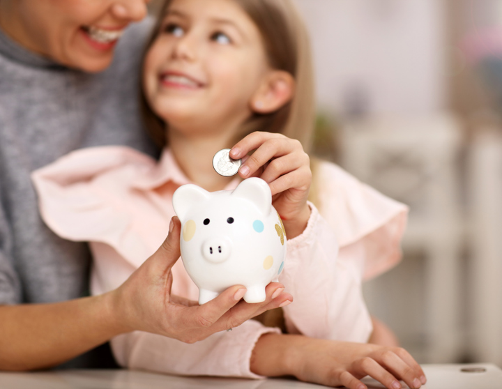Ein kleines Mädchen sitzt bei ihrer Mama auf dem Schoß während die Mama ein Sparschwein in der Hand hält und das kleine Mädchen einen Euro in das Sparschwein tut