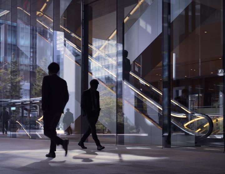 Two men in suits walk past each other in front of a modern glass building.