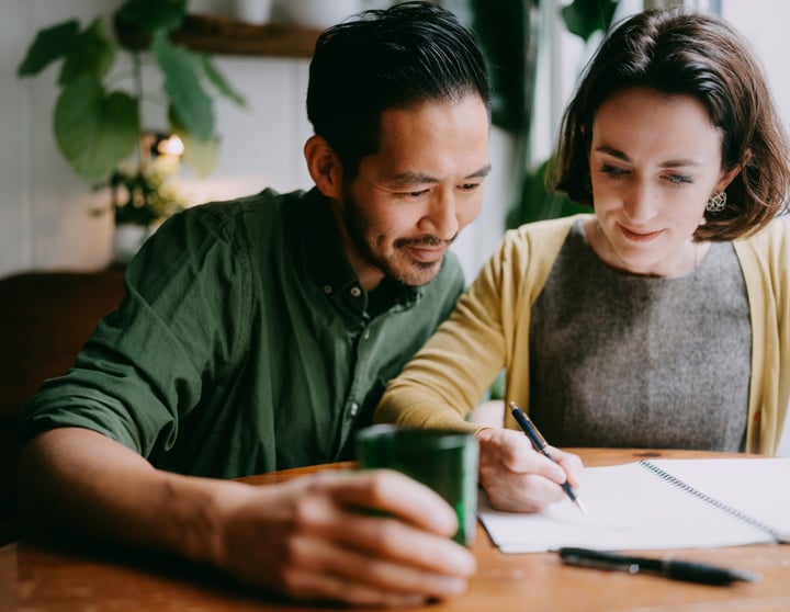 Ein brünetter Mann und eine brünette Frau gehen gemeinsam durch ihren Finanzplaner, um einen Überblick über die eigenen Finanzen zu bekommen