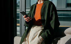 Frau in dunkelgrünem Mantel und rotem Pullover mit einem Smartphone in der Hand.