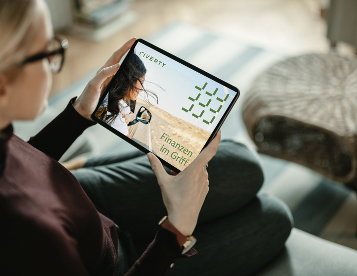 Eine blonde Frau sitzt auf dem Sofa und schaut sich auf ihrem Tablet den Riverty Finanzplaner.