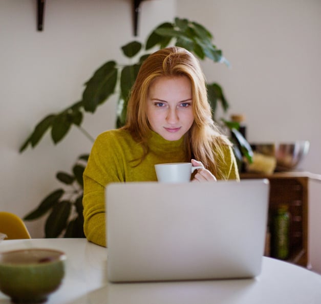 En kvinde sidder ved et bord med en kaffekop i hånden og ser koncentreret på sin computer