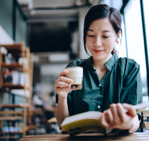 Eine leicht lächelnde Frau, sitzend in einem Cafe mit Buch in der einen und Kaffee in der anderen Hand