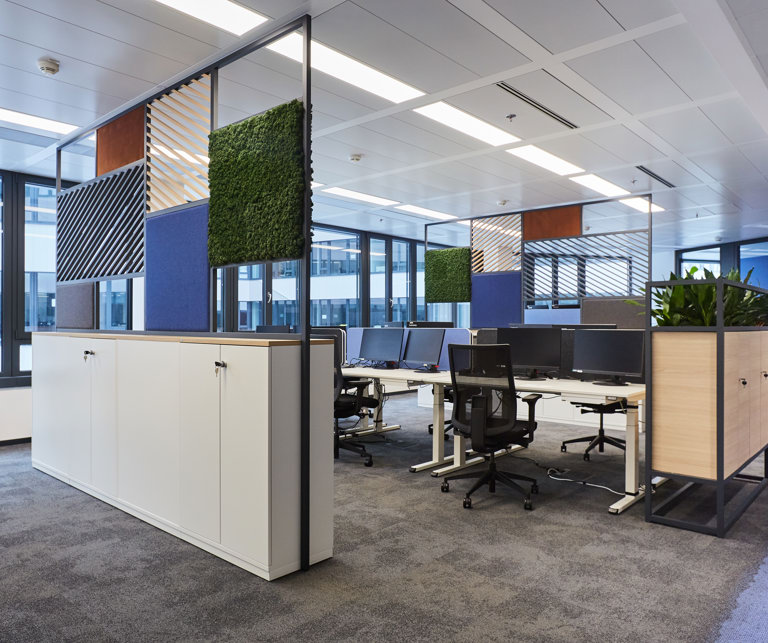Modernes Büro mit offener Raumgestaltung, skandinavischen Designelementen, Pflanzen und Raumtrennern. Jeder Schreibtisch ist mit zwei Bildschirmen ausgestattet.