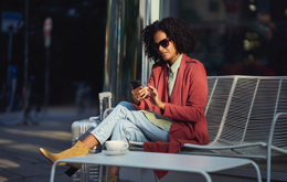 nainen istuu ulkona nauttien kahvista ja käyttäen puhelinta