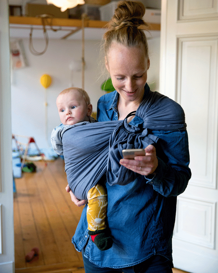 En kvinna bär ett barn i en sjal och tittar i sin mobil