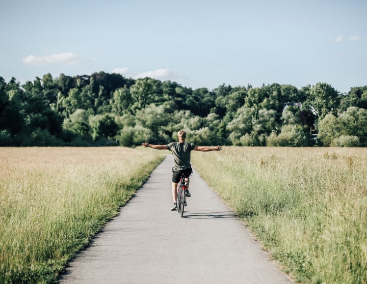 Ein Mann in einem grünen T-Shirt fährt mittig auf einer Straße Fahrrad und streckt dabei seine Arme von sich. Rechts und links von ihm befinden sich grüne Wiesen