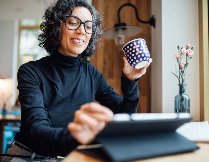 Eine brünette Frau sitzt am Tisch, hält eine blauweiß gepunktete Tasse in der Hand und tippt auf ihrem Tablet