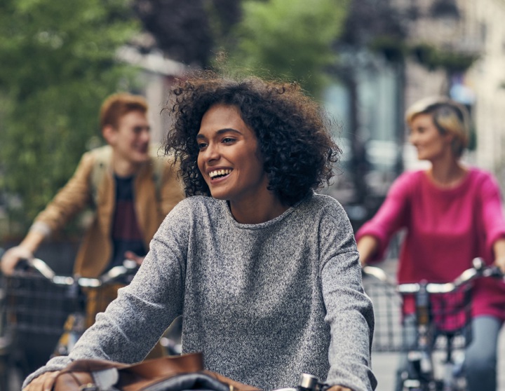 Im Vordergrund fährt eine brünette Frau lächelnd auf ihrem Fahrrad. Im Hintergrund fahren eine blonde Frau und ein Mann mit roten Haaren auf ihren Fahrrädern und unterhalten sich