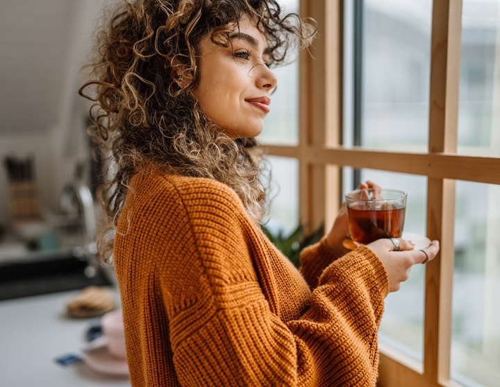 Eine Frau mit brünetten Locken trägt einen orangenen Pullover, schaut aus dem Fenster und hält eine Tasse Tee