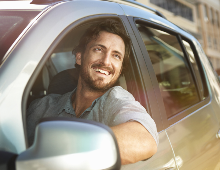 Ein brünetter Mann sitzt lächelnd in einem silbernen Auto und lehnt sich leicht aus dem offenen Fenster