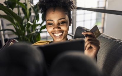 Eine junge brünette Frau sitzt auf dem Sofa, hält eine Kreditkarte in der Hand und shoppt auf ihrem Tablet die besten Black Friday Deals