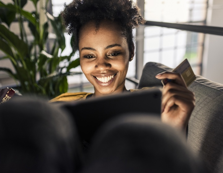 Eine brünette Frau schaut lächelnd auf ihr Tablet und hält dabei eine Kreditkarte in ihrer Hand