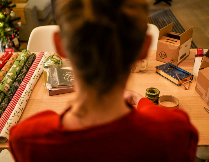 Eine junge Frau sitzt vor einem Tisch, der voll mit buntem Geschenkpapier und Weihnachtskarten ist