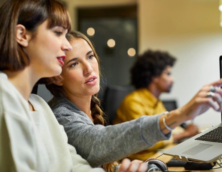 Zwei junge Frauen sitzen vor einem PC-Bildschirm. Die eine zeigt auf etwas. 