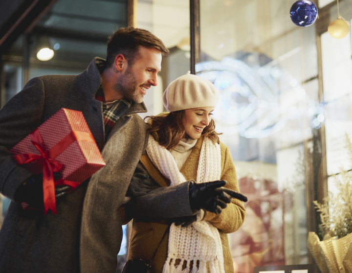Ein Mann hält ein rot verpacktes Geschenk und eine Frau mit Mütze stehen vor einem Schaufenster und schauen rein
