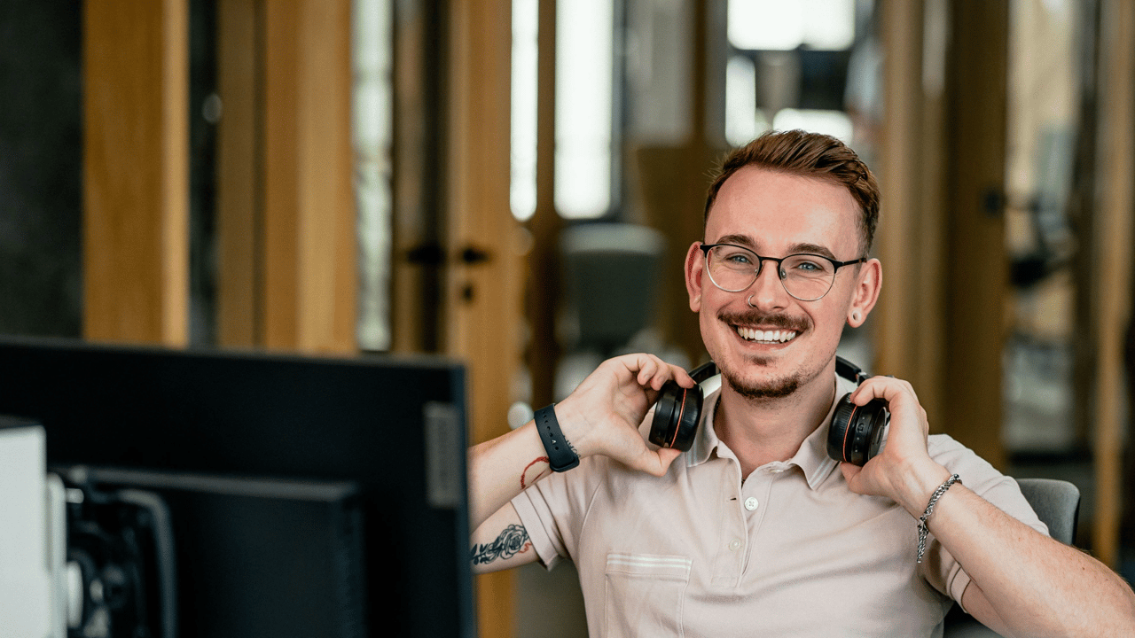 Lächelnder junger Mann mit Brille und kurzen braunen Haaren sitzt an einem Schreibtisch in einem modernen Büro und trägt schwarze Kopfhörer um den Hals. Er trägt ein beigefarbenes Poloshirt und eine Smartwatch. Im Vordergrund ist ein Computerbildschirm zu sehen, dahinter sanft verschwommen ein warmes Interieur aus Holz.