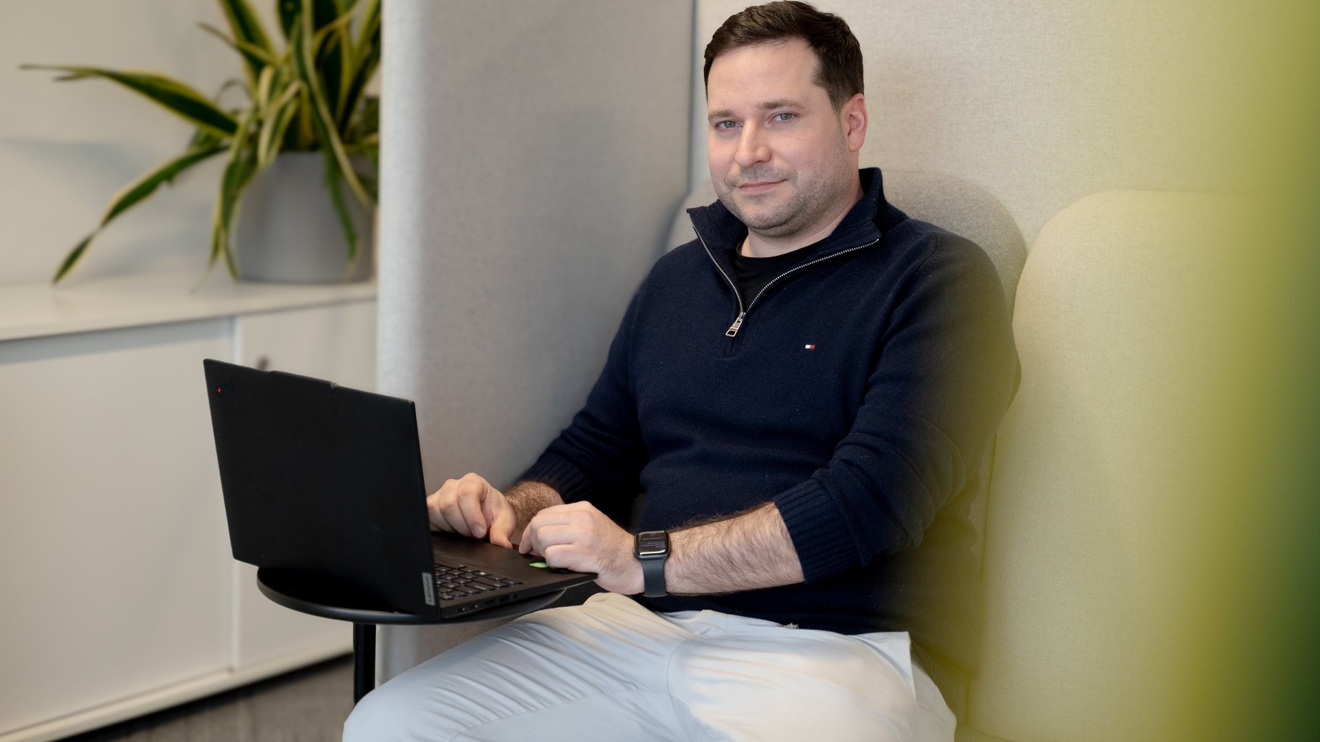 Ein Mann sitzt in einer gepolsterten Nische und arbeitet an einem Laptop auf einem kleinen Tisch. Er trägt einen dunklen Pullover und helle Hose, hat eine Uhr am Handgelenk und blickt in die Kamera. Im Hintergrund steht eine Zimmerpflanze auf einem Sideboard.