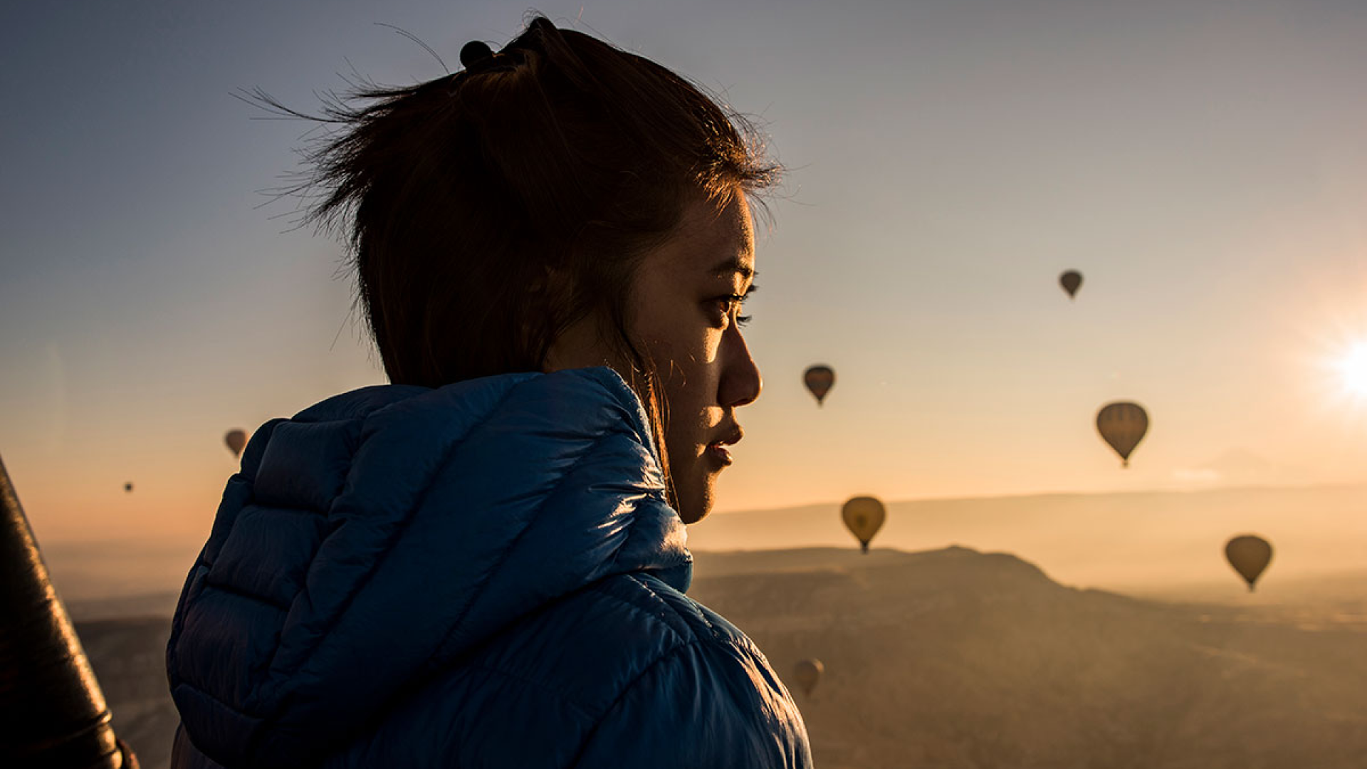 Frau, die bei Sonnenuntergang auf einem Berg steht und in die Ferne schaut, hinter ihr sieht man mehrere Heißluftballons.