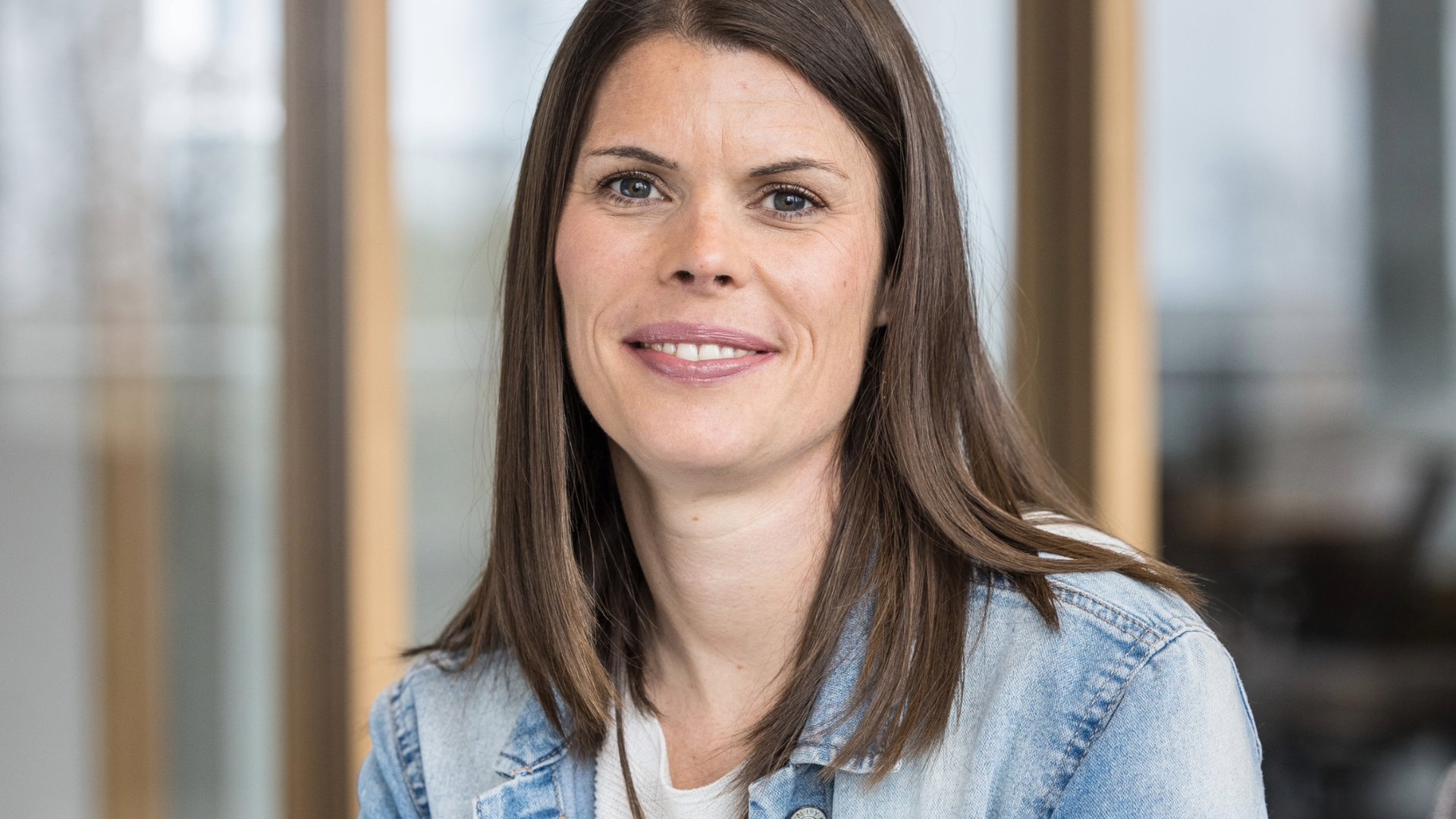 Circular portrait photo of a person with long brown hair and a friendly smile. Denim jacket, light background.