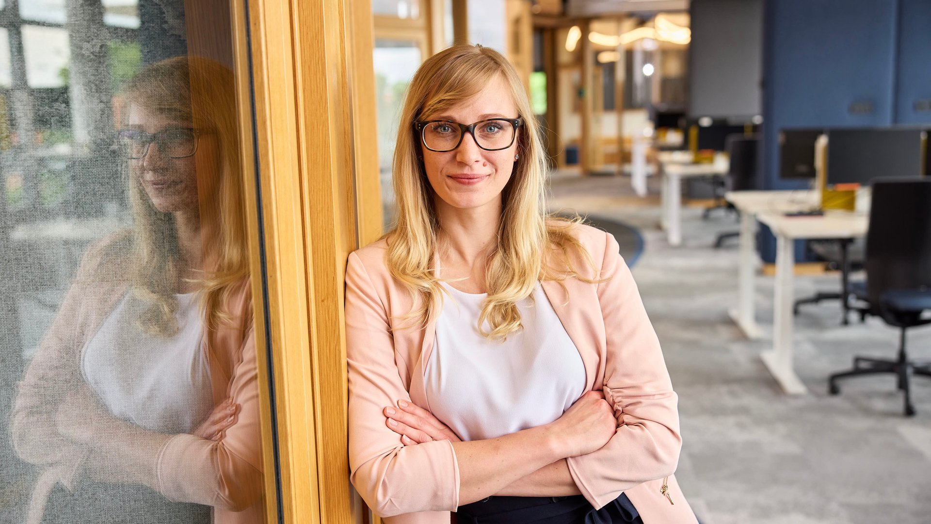 Regina im Büro lächelt in die Kamera und lehnt sich mit verschränkten Armen an eine Wand an