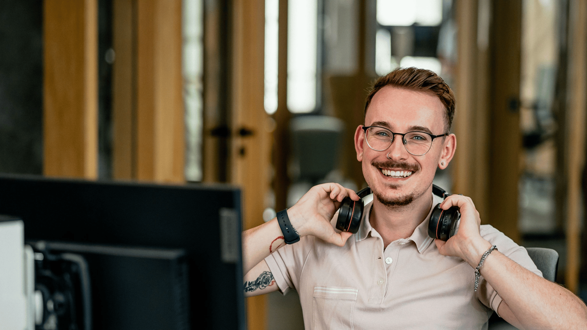 Smiling young man with glasses and short brown hair sits at a desk in a modern office, holding black headphones around his neck. He wears a beige polo shirt and a smartwatch, with a computer monitor in the foreground and warm wooden interiors softly blurred behind him.