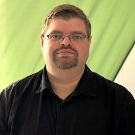 Eine Person steht vor einem halbtransparenten, grün-weißen Hintergrund, der an ein modernes Stoffbanner oder Segel erinnert. Die Person trägt ein schwarzes Hemd mit Kragen und eine Brille mit schmalem Metallrahmen. Sie hat kurzes, braunes Haar, einen gepflegten Kinnbart und schaut mit ruhigem, neutralem Gesichtsausdruck direkt in die Kamera. Das Licht fällt von der linken Seite ein und erzeugt weiche Schatten. Es sind keine Marken, technischen Geräte oder barrierebezogenen Symbole sichtbar.