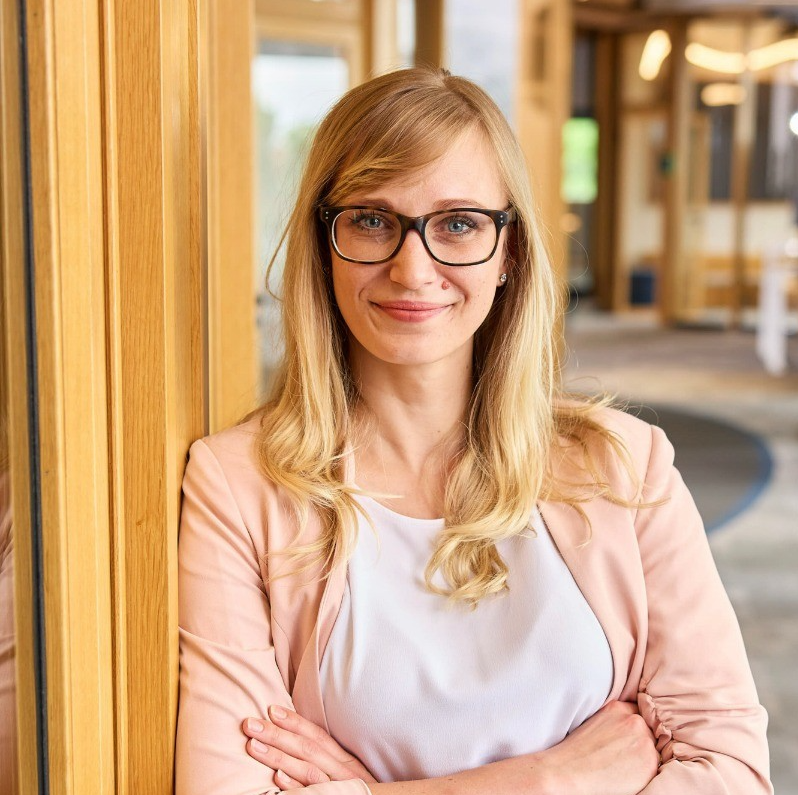 blonde woman with glasses, wearing a light pink blazer and white top, leans with folded arms against a wooden doorframe, with her reflection visible on a adjacent glass panel in a modern office space.