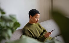 Frau sitzt auf einem Sofa in einem modernen Wohnzimmer und schaut auf ihr Smartphone, umgeben von grünen Pflanzen bei weichem Tageslicht.